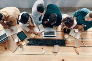 A top-down view of a team collaborating around a wooden table with laptops, notebooks, and tablets, working together on a project in a modern tech business environment.