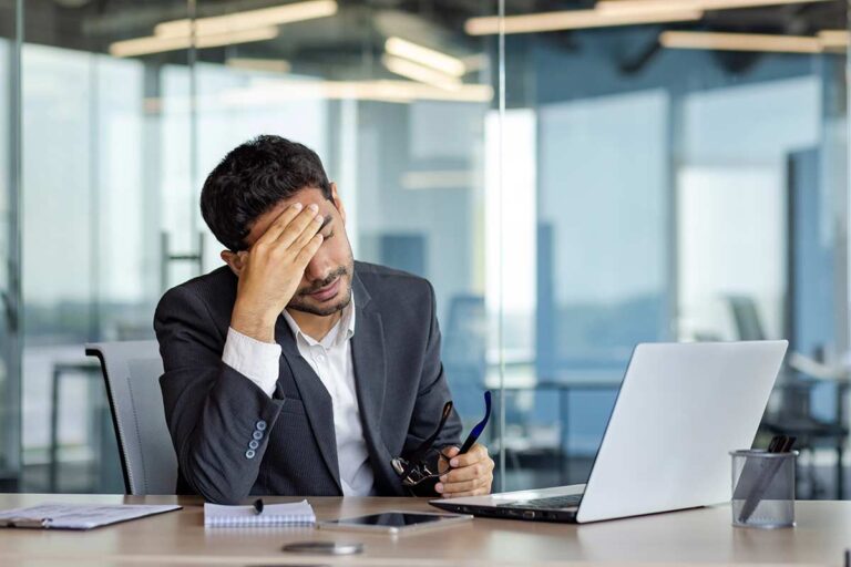 An overwhelmed executive in a modern office holding his head while working on a laptop, with notes and glasses on the desk.