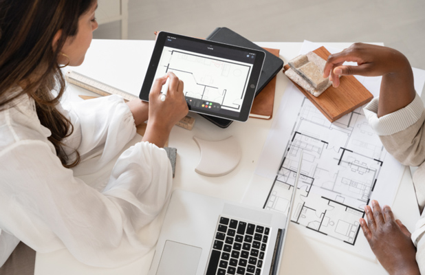 Two women working on a design project, using architectural blueprints and a tablet in a modern workspace, emphasizing collaboration and innovation.