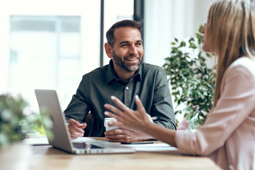 Professional consultant in dark green shirt smiling during meeting with client, discussing project details over laptop in bright modern office with indoor plants