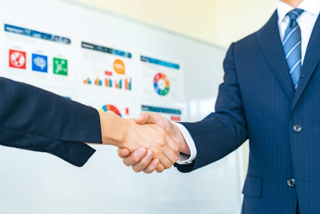 Two business professionals shaking hands in front of a whiteboard displaying charts and graphs, symbolizing a successful merger agreement and strategic planning.