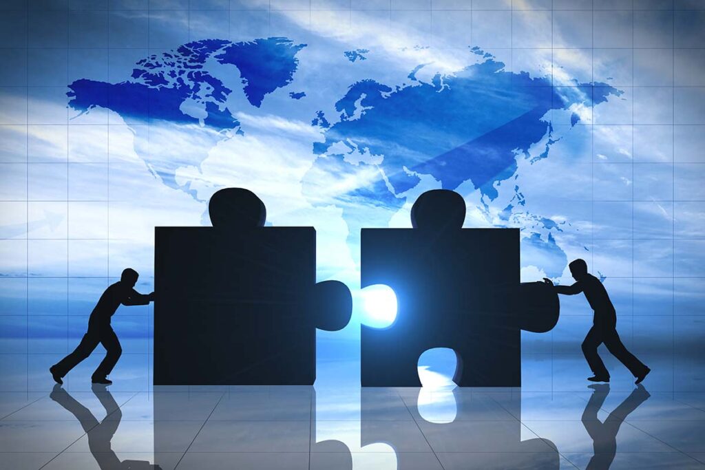 Silhouettes of two people pushing large puzzle pieces together, symbolizing business mergers and collaboration, with a world map in the background.